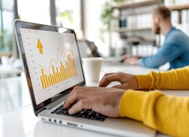 Person in yellow sweater working on a laptop analyzing financial data and graphs in a modern office setting with another person in background. photo