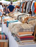 A variety of colorful woven blankets displayed on a market stall, with people browsing in the background under sunny weather. photo