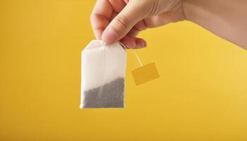 Hand holding a tea bag against a solid yellow background. Perfect for concepts related to tea, beverages, and relaxation. photo