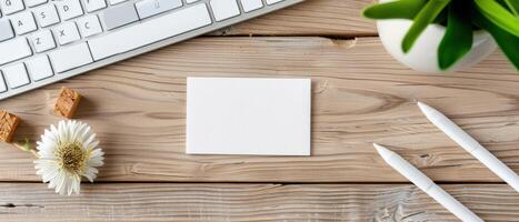 Top view of a clean desktop with blank white card, keyboard, pens, plant, and flower. Ideal for workspace and stationery themes. photo