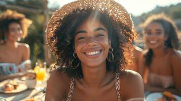 A cheerful young woman with curly hair and a hat, smiling joyfully at an outdoor summer gathering with friends. photo