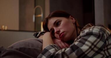 A calm and bored brunette girl in a checkered shirt leans on a pillow on a gray sofa she is bored and looks away in a modern apartment in the evening video