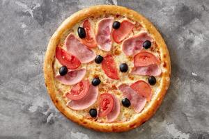 Thin classic pizza with melted cheese, ham, tomatoes and black olives on gray stone surface photo