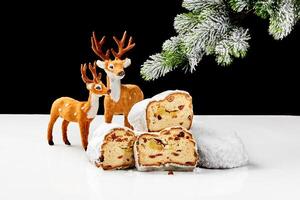 Slices of sweet stollen with nuts and dried fruit on table with toy deers and snow-covered spruce branch photo