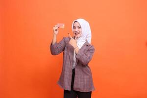 Young cheerful Asian woman hijab suit holding plastic debit credit card while pointing at it isolated on orange background. satisfied with service for financial concept, mobile transactions, payments photo