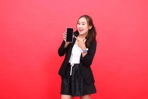 gesture of cheerful mature Asian career woman wearing a black formal suit holding a smartphone while pointing at it, for technology, business, advertising, financial content photo