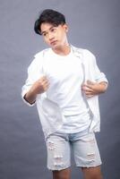 cool asian man posing standing head tilted left forward in front of the camera while holding his shirt in both hands indoors. fashion promotion man shirt photo session in the gray background
