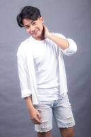 elegant asian man posing standing sideways in front of the camera while holding his left cheek indoors. fashion promotion man shirt photo session in the gray background studio