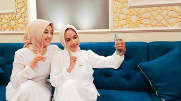 portrait of a pair of beautiful Asian women sitting on the sofa smiling cheerfully posing for a selfie in front of a cellphone with an elegant Arabic nuance in the background of Islamic calligraphy photo