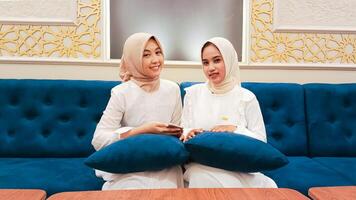 two beautiful Asian women sitting on the sofa smiling cheerfully holding cellphone gadgets with an elegant Arabic nuance of Islamic ornament calligraphy in the background photo