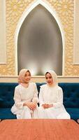portrait of a couple of women sitting on a sofa smiling cheerfully with a welcome hand gestur facing the camera with an elegant Arabic Islamic calligraphy background photo