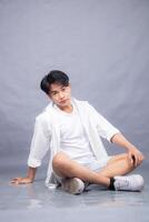 elegant asian man posing sitting in front of the camera while his hands hold his knees indoors. fashion promotion man shirt photo session in the gray background studio