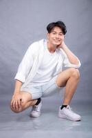 elegant smiling asian man posing in upright squats in front of the camera while holding his hands to his cheeks indoors. fashion promotion man shirt photo session in the gray background studio
