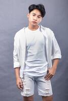 elegant asian man posing standing with his head straight forward in front of the camera while holding his pants indoors with both hands. fashion promotion man shirt photo session in the gray