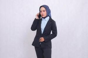 A young Asian office executive woman wearing a hijab stands elegantly while calling someone on her cellphone. Beautiful Muslim woman in blue shirt used for business, finance and technology content photo