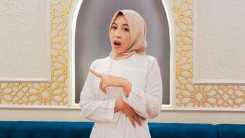 The charm of an Asian woman wearing a hijab looks at the camera with a shocked expression, pointing her hand to the right with an Islamic ornamental calligraphy background photo
