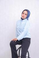 young Asian office woman wearing a hijab, blue shirt, sitting in a chair, smiling elegantly, hands on thighs. Beautiful Muslim women use business, finance and fashion content photo