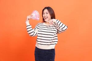 an Asian woman with a cheerful expression at the camera stands holding several rupiah bills while pointing at them. for fashion, business and finance concept photo