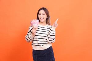 an Asian girl with a happy expression at the camera stands holding several rupiah bills while pointing up. for fashion, business and finance concept photo