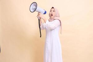 woman asian mature wearing hijab slanted to the right candidly shouting loudly, holding a loudspeaker megaphone with both hands. Technology, broadcast and promotion concept photo