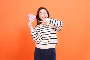 an Asian woman with a cheerful expression, candid wink, standing while holding a number of rupiah bills while pointing at them. for fashion, business and finance concept photo