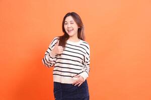 Portrait of a beautiful cheerful confident asian girl winking standing with hands to waist gesture cool okay thumbs up and looking at camera isolated on orange background wearing white sweater photo