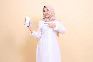 Young woman indonesia muslim wearing a hijab smiles cheerfully holding a smartphone gadget to the right in her hand while pointing at it. Lifestyle, business and commercial concept photo
