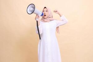 girl indonesia wearing hijab slanted to the right candidly shouts enthusiastically, holding a loudspeaker megaphone and clenching her fists upwards. Technology, broadcast and promotion concept photo
