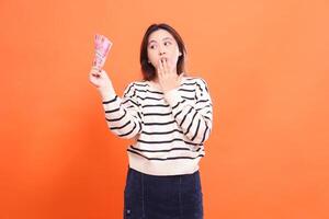 an Asian girl with a shocked expression stands while holding a number of rupiah notes while candid. for fashion, business and finance concept photo