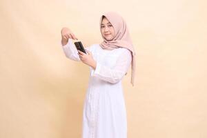 Asian Muslim woman wearing a hijab smiles at the camera, operating a cellphone gadget with both hands while pointing. Lifestyle, technology and promotion concept photo