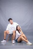 smiling man and woman posing sitting in front of camera holding ladder holding arms back indoors. fashion promotion couple photo session in gray background studio