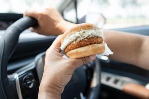 Asian lady holding hamburger to eat in car, dangerous and risk an accident. photo