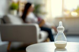 Baby feeding bottle with milk on a table in a bright room, in the background, out of focus, a mother holds a newborn in her arms while sitting on the sofa photo
