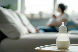 Baby feeding bottle with milk on a table in a bright room, in the background, out of focus, a mother holds a newborn in her arms while sitting on the sofa photo