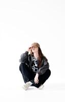 Young woman in a bomber jacket, jeans and a cap with braces poses while sitting in the studio, on a white background photo