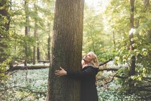 Blonde woman in a long black coat smiles and hugs a massive tree in the wilderness, symbolizing a deep connection and commitment to protecting our most vulnerable natural environment photo