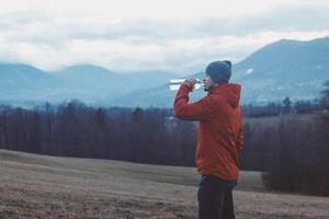 Adventurer on a hike drinks clear water from a glass bottle in a meadow in front of the mountains during a break. Refreshment break photo
