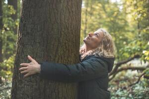 Blonde woman in a long black coat smiles and hugs a massive tree in the wilderness, symbolizing a deep connection and commitment to protecting our most vulnerable natural environment photo