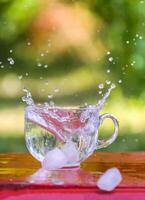 Splash of cool fresh water with ice cube in transparent glass cup in the table outoors in summer day. photo