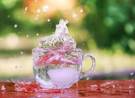 Splash of cool fresh water with ice cube in transparent glass cup in the table outoors in summer day. photo