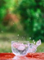 Splash of cool fresh water with ice cube in transparent glass cup in the table outoors in summer day. photo