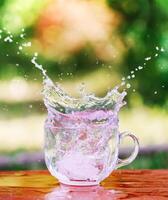 Splash of cool fresh water with ice cube in transparent glass cup in the table outoors in summer day. photo