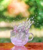 Splash of cool fresh water with ice cube in transparent glass cup in the table outoors in summer day. photo