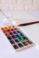 Watercolor paints, brushes, water in glass and paper on white background. Flat lay, top view photo