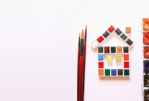 Watercolor paints in the shape of a house, brushes and paper on white background. Flat lay, top view photo