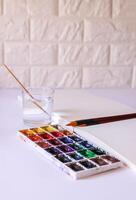 Watercolor paints, brushes, water in glass and paper on white background. Flat lay, top view photo