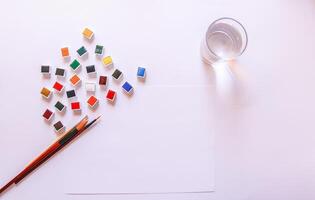 Watercolor paints, brushes, water in glass and paper on white background. Flat lay, top view photo