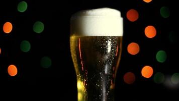 A glass with a cold light foamy beer stands on a table on a black background with colored lights. Drops of water flow down the glass. video