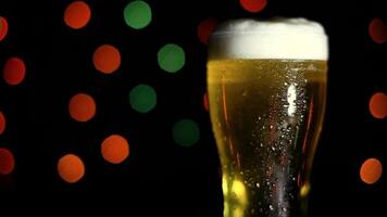 A glass with a cold light foamy beer stands on a table on a black background with colored lights. HD video