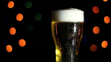 A glass with a cold light foamy beer stands on a table on a black background with colored lights. Drops of water flow down the glass. Mood of the holiday video